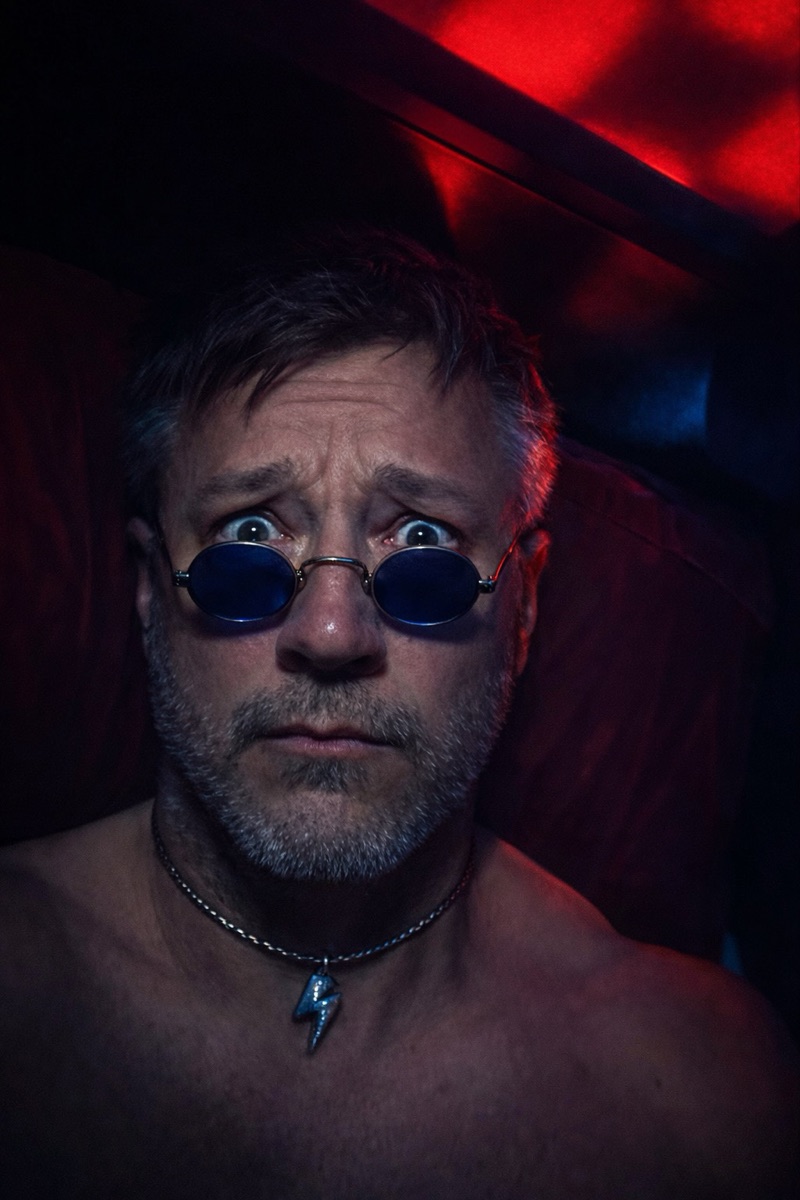 Fugdale lit hard in red, bare-shouldered in small round blue-tinted glasses, caught mid-alarm.