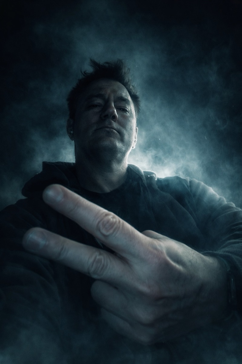 Fugdale in smoke and low blue light, head down, throwing a two-finger peace sign with a tattooed hand.