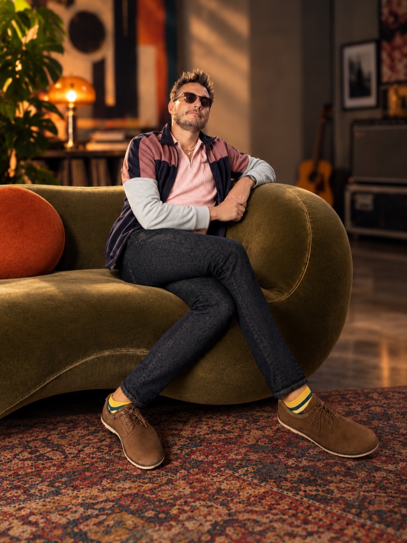 Fugdale reclined on a curved olive velvet sofa in a warmly lit room, sunglasses on, color-blocked jacket, patterned jeans and suede shoes.